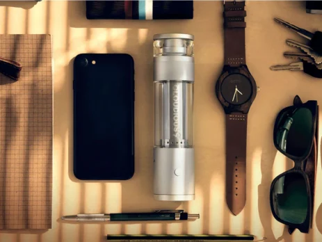 A Hydrology9 Vaporizer is arranged among other daily use items on a table, including sunglasses, a cellphone and watch.