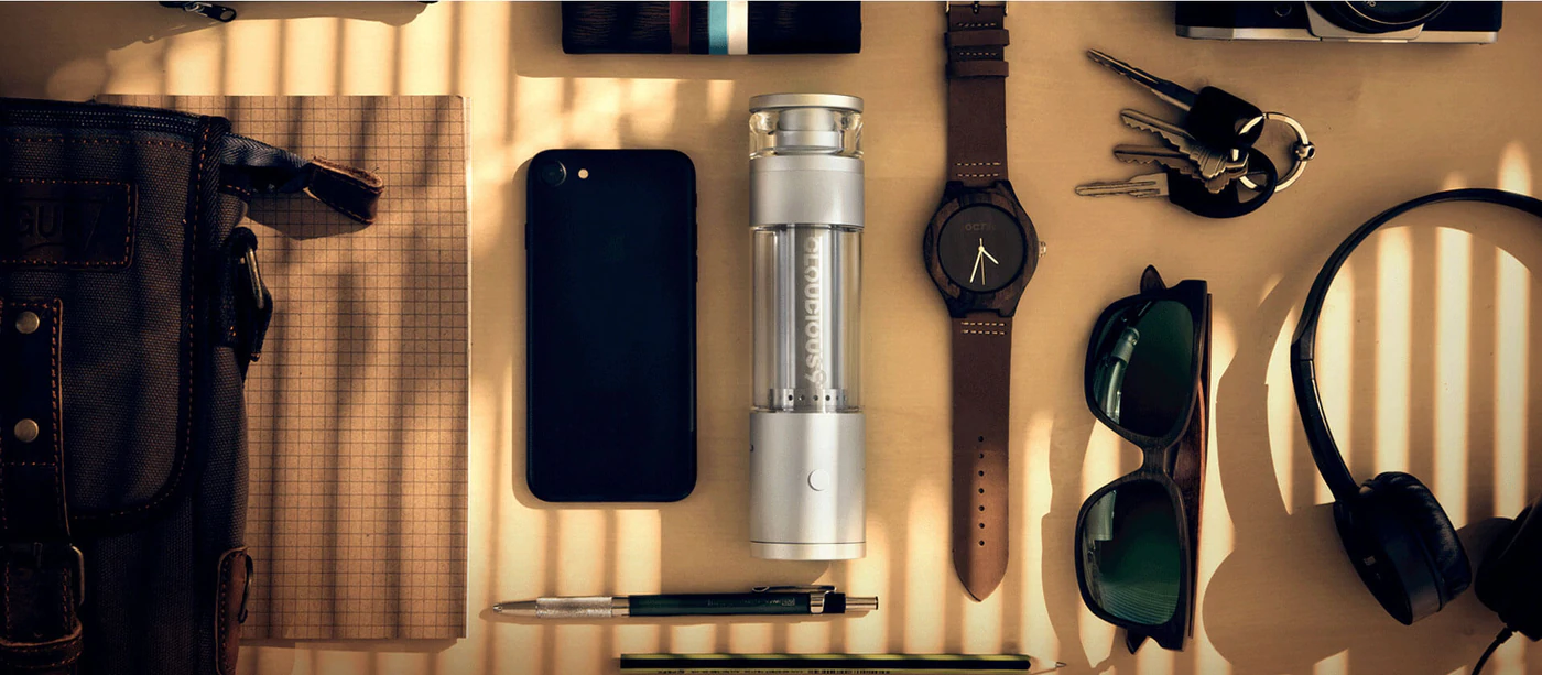 A Hydrology9 Vaporizer is arranged among other daily use items on a table, including sunglasses, a cellphone and watch.